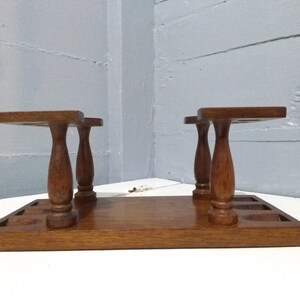 May include: Wooden pipe rack with six slots for pipes. The rack has a brown finish and is made of wood. It has a rectangular base and two sets of three turned legs.