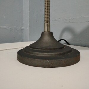 May include: Close-up of a dark gray desk lamp base with a round, tiered design. The lamp has a flexible, metallic neck and a power cord. The base is made of a heavy, possibly cast iron material, and sits on a white surface.