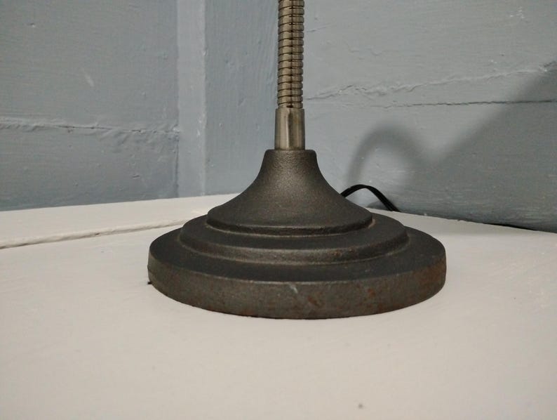 May include: Close-up of a dark gray desk lamp base with a round, tiered design. The lamp has a flexible, metallic neck and a power cord. The base is made of a heavy, possibly cast iron material, and sits on a white surface.