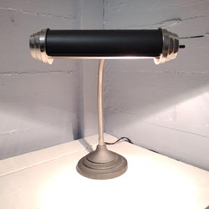 May include: A vintage-style desk lamp with a black cylindrical shade and a flexible, silver-colored gooseneck. The lamp has a silver-colored, decorative trim and a round base. The lamp is plugged into an electrical outlet.