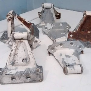 May include: Five vintage metal hinges, some painted white and some with rust. The hinges are in various sizes and shapes, with some having a triangular design.