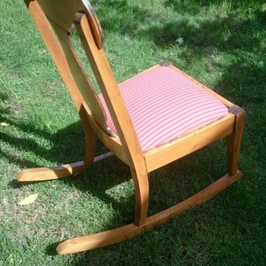 Puede incluir: Una mecedora de madera con un coj&iacute;n a rayas rosa y blanco. La silla tiene un respaldo alto y un asiento curvo.
