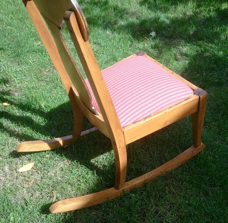 Puede incluir: Una mecedora de madera con un coj&iacute;n a rayas rosa y blanco. La silla tiene un respaldo alto y un asiento curvo.