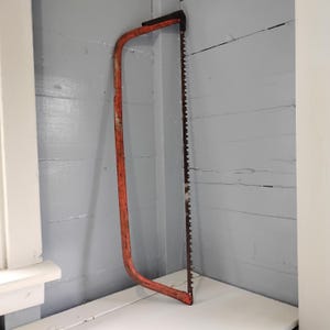 May include: A vintage bow saw featuring a bright orange metal frame and a long, straight steel blade with sharp, jagged teeth. The tool has a simple, functional design with a sturdy handle integrated into the frame, showing signs of wear.