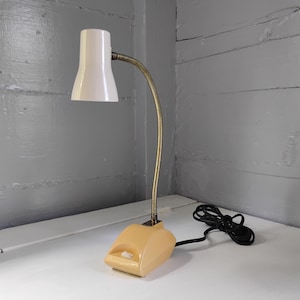 May include: A desk lamp featuring a white conical shade, a flexible brass-toned gooseneck arm, and a cream-colored plastic base with a white power switch. A black electrical cord with a plug is attached to the rear of the base unit.