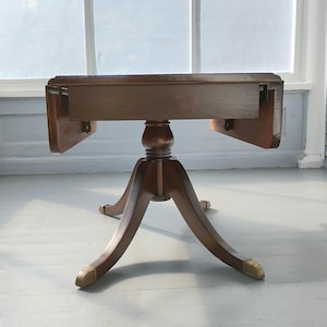 May include: A dark brown, antique drop-leaf table with a pedestal base and brass-capped feet. The table's top is rectangular with two drop leaves. The table is approximately 30 inches long when the leaves are down.