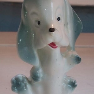 May include: A white ceramic figurine of a dog with blue ears and a pink tongue. The dog is sitting with its legs tucked under its body.