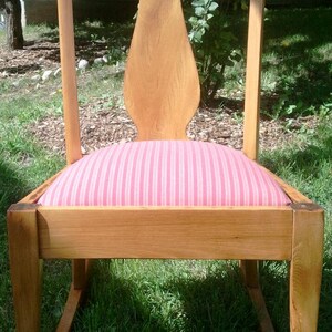 Puede incluir: Una silla mecedora de madera con un coj&iacute;n a rayas rosa y blanco. La silla tiene un respaldo alto y un asiento curvo.