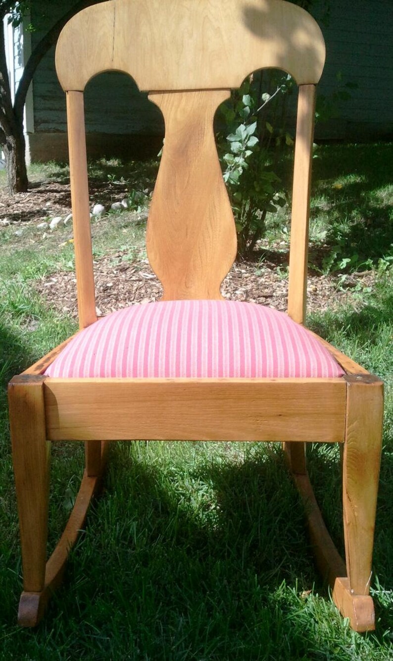 Puede incluir: Una silla mecedora de madera con un coj&iacute;n a rayas rosa y blanco. La silla tiene un respaldo alto y un asiento curvo.