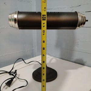 May include: A black desk lamp with a cylindrical shade and a round base. The lamp is approximately 13 inches tall, with silver-colored accents on the ends of the shade. A yellow measuring tape is placed next to the lamp.