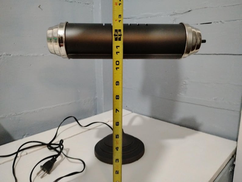 May include: A black desk lamp with a cylindrical shade and a round base. The lamp is approximately 13 inches tall, with silver-colored accents on the ends of the shade. A yellow measuring tape is placed next to the lamp.