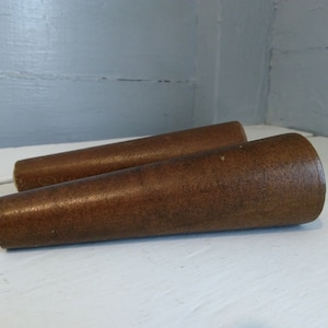 May include: Two brown wooden furniture legs with a tapered design. The legs are a warm brown color and appear to be made of solid wood. They are cylindrical with a slightly wider top and a narrower bottom.