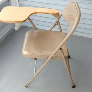 May include: A beige metal folding chair with a small wooden desk attached to the right armrest. The chair is in a room with a light gray floor and white walls.