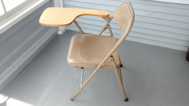 May include: A beige metal folding chair with a small wooden desk attached to the right armrest. The chair is in a room with a light gray floor and white walls.