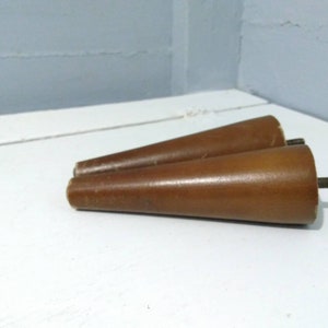 May include: Two brown wooden furniture legs with a tapered design. Each leg has a threaded metal rod extending from the base for attachment. The legs are made of a light brown wood and are set against a white surface.