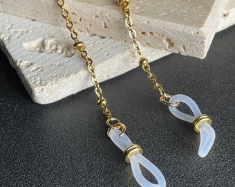 May include: A gold chain with small round beads and two clear silicone loops at the ends. The chain is on a light brown surface.