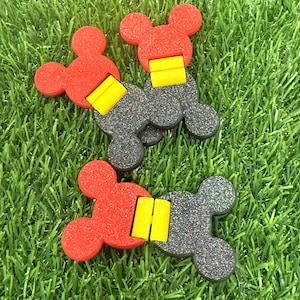 May include: Three Mickey Mouse-shaped accessories. Each has a red head, a yellow rectangular piece, and a dark gray body with glitter. The accessories are arranged on a bed of green grass.