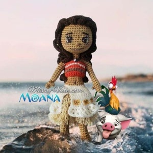 May include: Crocheted Moana doll with a brown and white skirt, a red and orange top, and brown hair. She is standing on a rock with a rooster and a pig beside her. The text "Missy-Ville.com MOANA" is visible in the background.