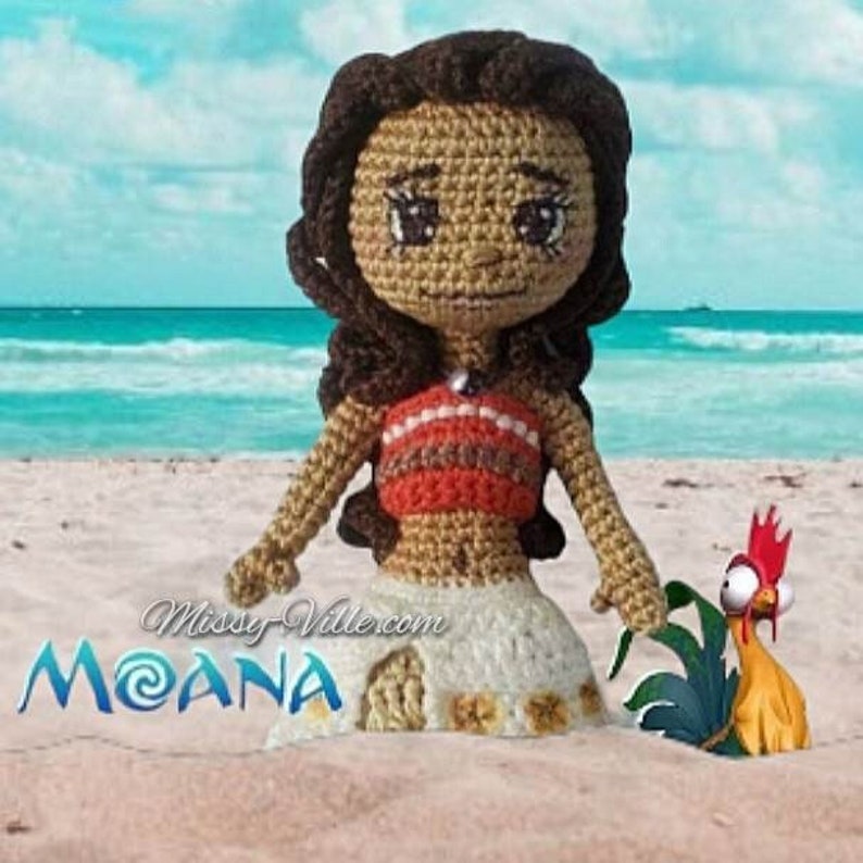 May include: A crocheted doll of Moana from the Disney movie Moana. The doll is wearing a red and brown top and a white skirt. The doll is standing on a beach with a rooster figurine beside it. The text "Moana" is visible in the image.