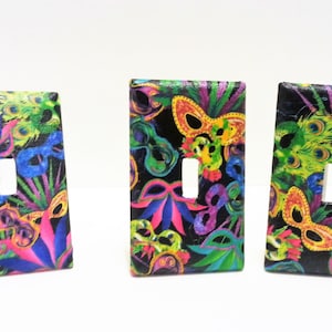 May include: Three light switch covers with a Mardi Gras theme. The covers are black with a colorful pattern of feathers, masks, and beads.
