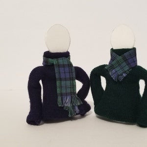 May include: Two sock puppets wearing sweaters and scarves. The puppet on the left is wearing a dark purple sweater and a green and blue plaid scarf. The puppet on the right is wearing a dark green sweater and a green and blue plaid scarf.