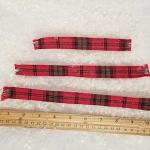 May include: Three red plaid ribbons with frayed edges are displayed next to a wooden ruler. The ribbons vary in length, with the longest measuring approximately 11 inches. The plaid pattern includes black and brown lines.