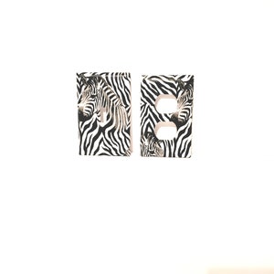 May include: Two white light switch covers with a zebra print design. The zebra print is black and white.