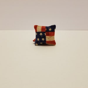 May include: Small, square pillow with a patriotic American flag design. The pillow features a blue field with white stars, and red and white stripes. The pillow is made of fabric and has a soft appearance.