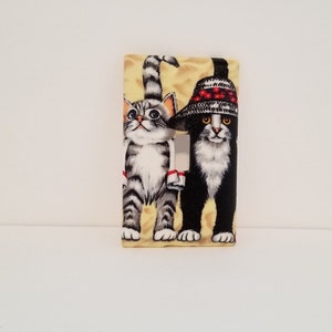 May include: A light switch cover featuring a black and white cat wearing a red and white hat, standing next to a gray and white tabby cat. The background is a yellow and white striped pattern.