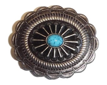 May include: A silver-toned metal belt buckle with a turquoise stone in the center. The buckle is decorated with a floral design and a scalloped edge.