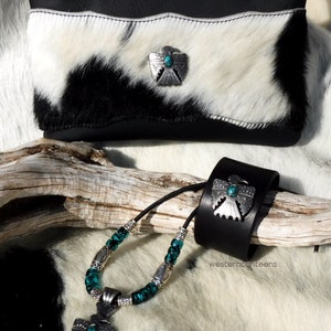May include: A black leather wrist cuff with a turquoise and silver eagle design. A matching necklace with turquoise and silver beads and a pendant with a turquoise and silver eagle design. The jewelry is on a white and black cowhide background.