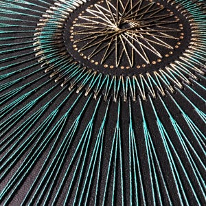 Op de afbeelding: Een close-up van een cirkelvormig borduurmotief op zwarte stof. Het ontwerp heeft een centrale stervorm met gouden draad en een omringende ring van turquoise draad.