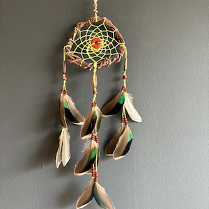 May include: A handcrafted dreamcatcher with a woven brown and yellow web, featuring a red center bead. The dreamcatcher is adorned with brown, green, and white feathers, and wooden beads. It hangs from a yellow string, perfect for home decor.
