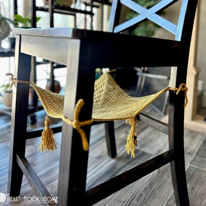 Może przedstawiać: Hamak dla kota wykonany na szydełku, w kolorze musztardowo-żółtym, wiszący na oparciu drewnianego krzesła. Hamak ma frędzle na końcach i jest przywiązany do krzesła żółtą sznurówką.