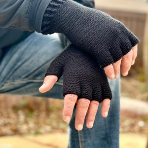May include: A pair of black fingerless gloves with a crocheted texture. The gloves are being worn by someone wearing blue jeans.