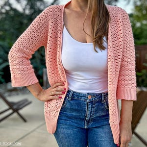 May include: A light peach crochet cardigan with a diamond pattern. The open-front sweater has three-quarter sleeves and a ribbed hem and cuffs. The cardigan is worn over a white vest top and blue jeans.