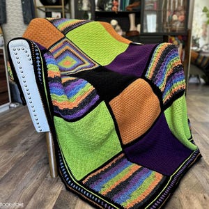 Peut inclure: Une couverture crochetée à la main aux couleurs vives, avec un motif patchwork de carrés noirs, orange, verts et violets. La couverture est drapée sur une chaise. Les bords sont bordés d'une bordure noire et arc-en-ciel. Un accessoire de maison douillet.