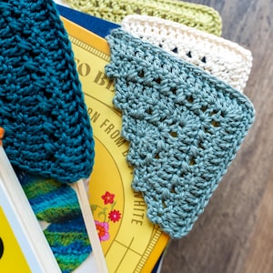 May include: Close-up of several handmade crochet bookmarks in various colors, including orange, teal, and green. The bookmarks are placed on top of a book with a yellow cover. The crochet work has a textured appearance.