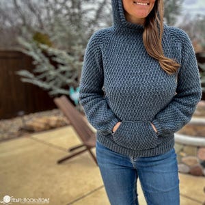 May include: A blue hooded sweater with a textured knit pattern and a front pocket. The sweater is being worn by a person with long brown hair.
