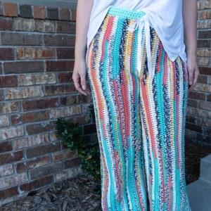 May include: A pair of wide-leg crochet trousers with a colourful striped pattern in shades of blue, pink, yellow, and white. The trousers have a drawstring waist and are made with a soft, lightweight yarn.