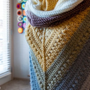 May include: A crocheted shawl with a gradient of blue, green, yellow, and white. The shawl is draped over a form and has a textured stitch pattern.