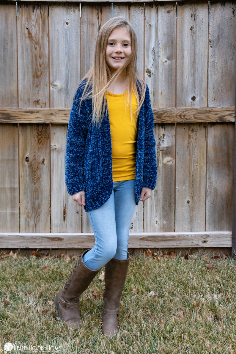 May include: A young person wearing a blue fuzzy cardigan over a yellow long-sleeved shirt, light blue jeans, and brown knee-high boots. The person is standing in front of a wooden fence.