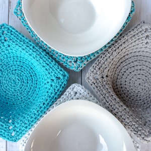 May include: Four crocheted pot holders in teal and gray, with white bowls on top. The pot holders are square and have a circular design in the center.
