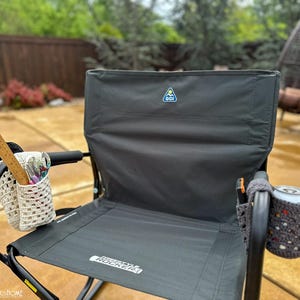 May include: A black folding chair with a gray fabric seat and back. The chair has a blue and yellow logo on the back that says "GCI Outdoor". The chair has a crocheted cup holder on the right armrest and a crocheted storage bag on the left armrest. The chair is sitting on a concrete patio.