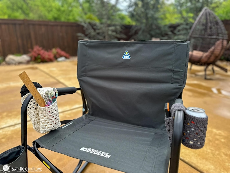 May include: A black folding chair with a gray fabric seat and back. The chair has a blue and yellow logo on the back that says "GCI Outdoor". The chair has a crocheted cup holder on the right armrest and a crocheted storage bag on the left armrest. The chair is sitting on a concrete patio.