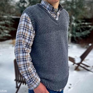May include: A gray knitted vest with a V-neck and a textured pattern. The vest is worn over a plaid shirt.