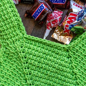 May include: A close-up of a bright green crocheted bag filled with various wrapped candies, including Snickers, Twix, and Tootsie Rolls. The bag has a textured weave pattern and a handle. The candies are scattered around the bag.