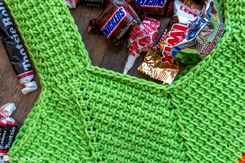May include: A close-up of a bright green crocheted bag filled with various wrapped candies, including Snickers, Twix, and Tootsie Rolls. The bag has a textured weave pattern and a handle. The candies are scattered around the bag.
