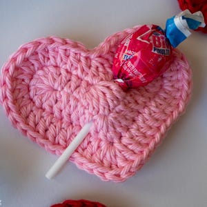 May include: A light pink crocheted heart-shaped holder featuring a central pocket that secures a red and blue wrapped lollipop. The lollipop stick extends from the bottom of the heart, which is crafted with a visible yarn stitch pattern.