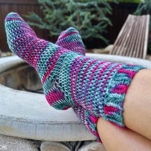 Könnte beinhalten: Gestrickte Socken in Türkis, Pink und Lila. Die Socken haben ein strukturiertes Muster und werden von jemandem getragen, der sich im Freien entspannt. Die Socken liegen auf einer Feuerstelle aus Beton.
